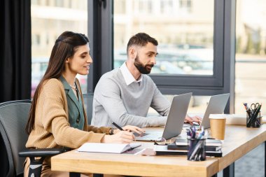 İki profesyonel bir ofiste bir masada oturup dizüstü bilgisayarlarıyla ilgilenerek modern bir şirket kültürü sergiliyorlar..