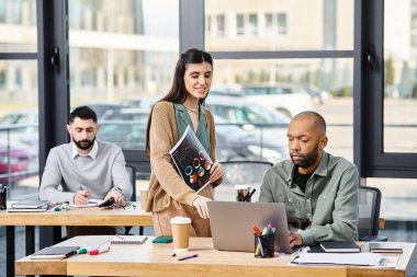 Hareketli bir ofiste dizüstü bilgisayarın önünde özgüvenle duran bir kadın, odaklanmış kararlılıkla bir projeyi denetliyor..