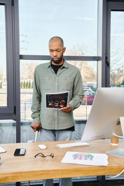 Bir adam güven içinde bir masanın önünde duruyor, elinde bir kitapla bir ofis ortamında verimli bir iş gününe hazırlanıyor..