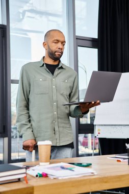 Bir adam dizüstü bilgisayarın önünde özgüvenle duruyor, modern ofis ortamında özenle bir proje üzerinde çalışıyor..