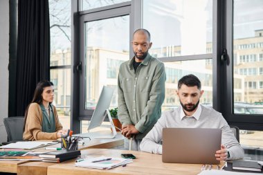 Masaya odaklanmış bir adam, yoğun bir ofis ortamında dizüstü bilgisayarla yazı yazıyor, bir iş projesinin kurumsal kültürüne dalmış..