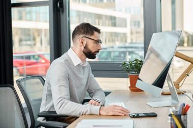 Bir adam masanın başında oturuyor, kendini bilgisayar ekranına kaptırmış, yoğun bir ofis ortamındaki bir projeye odaklanmış..