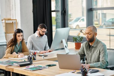 Diverse group of colleagues sitting around a table in an office, working intently on laptops together for a project. clipart