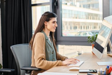 Masada çalışmaya dalmış bir kadın, canlı bir ofis ortamında bilgisayar kullanıyor..