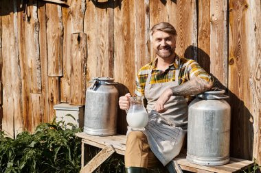 cheerful attractive farmer sitting near his house with jar and churns of milk and smiling at camera clipart