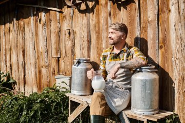 cheerful attractive farmer sitting near his house with jar and churns of milk and smiling away clipart