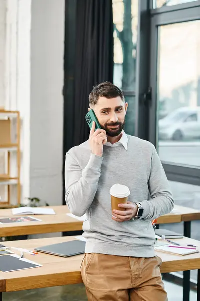 A man in a business setting holds a cup of coffee while talking on a cell phone.
