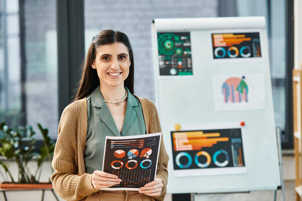 A woman stands by a whiteboard holding charts. She appears to be leading a presentation in a corporate office setting.