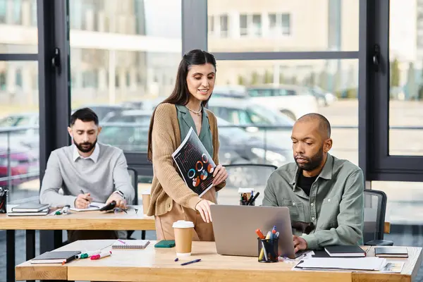 Hareketli bir ofiste dizüstü bilgisayarın önünde özgüvenle duran bir kadın, odaklanmış kararlılıkla bir projeyi denetliyor..