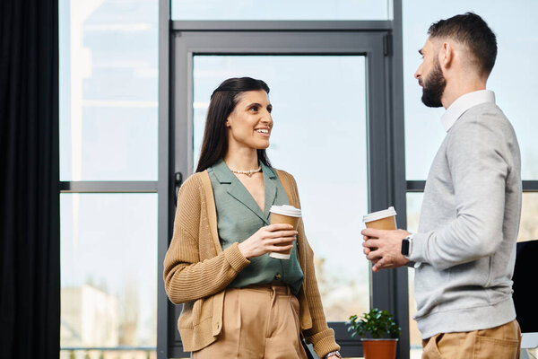 A man and woman standing, holding coffee cups, engaged in a conversation in a corporate office setting.
