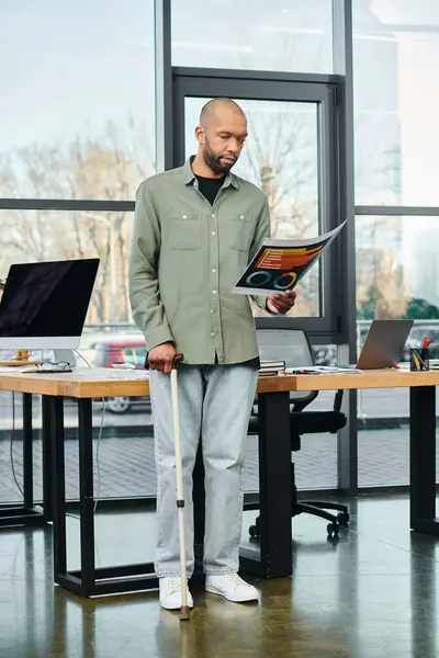 stock image disabled man stands with walking cane in front of a desk, holding charts in an office setting.