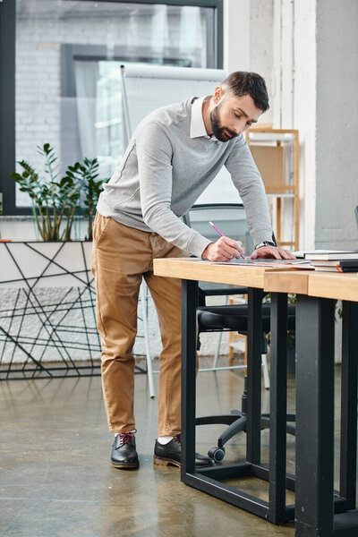A man focused on his laptop, standing at a desk in a busy office, working on a project integral to the corporate culture.