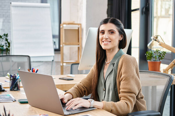 A professional woman with long hair sitting at a desk, focused on her laptop while working in an office setting.