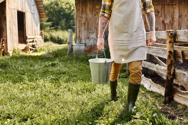 Çiftlikteyken elinde kovayla kollarında dövmeleri olan kendini işine adamış yetişkin bir adamın kesilmiş görüntüsü.