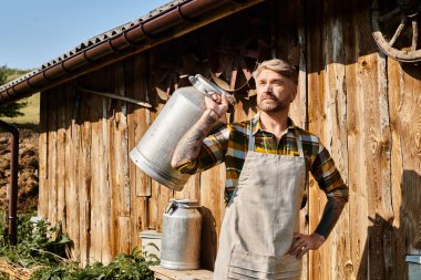 appealing bearded man in casual attire with tattoos posing with milk churns and looking away, farmer clipart