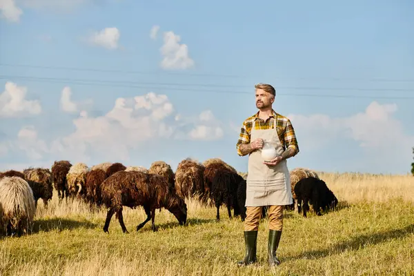 appealing modern farmer with tattoos and beard holding jar of fresh milk surrounded by sheeps