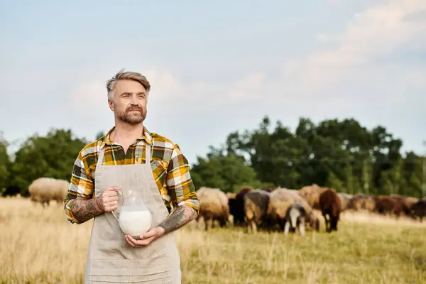 handsome bearded modern farmer with tattoos holding jar of fresh milk with sheeps on backdrop
