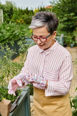 Neşeli, güzel, olgun bir kadın. Gözlüklü ve eldivenli. Bahçesinde poz veriyor ve başka tarafa bakıyor.