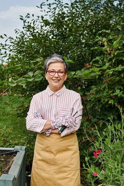 jolly mature woman in casual attire with glasses holding rakes for gardening and smiling at camera