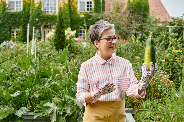 привлекательная веселая зрелая женщина в повседневной одежде в очках, держащих свежие цуккини в саду