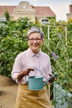 cheerful mature woman with glasses holding watering can and smiling at camera near house in England clipart