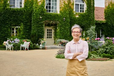 beautiful jolly mature woman with glasses in cozy attire looking at camera near her house in England clipart