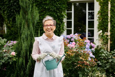 attractive mature jolly woman with glasses holding watering can and smiling happily at camera clipart