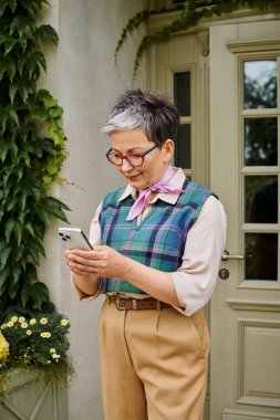 sofistike, olgun, gözlüklü bir kadın İngiltere 'deki evinin yanındaki telefonuna bakıyor.