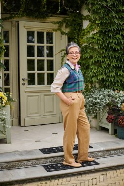 Güzel, güler yüzlü, gözlüklü, elleri ceplerinde poz veren ve kameraya gülümseyen bir kadın.