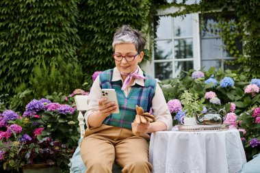 mature cheerful woman sitting in garden at tea time and looking at phone near house in England clipart