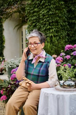 mature cheerful woman sitting in garden at tea time and talking by phone near house in England clipart