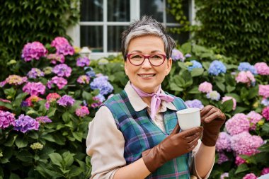 Şık giyinmiş zarif olgun bir kadın İngiltere 'de bir evin yakınında çay içiyor ve kameraya gülümsüyor.
