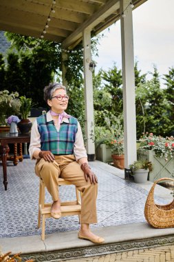 Moda düşkünü, olgun bir kadın İngiltere 'deki evinin verandasında oturuyor ve başka tarafa bakıyor.