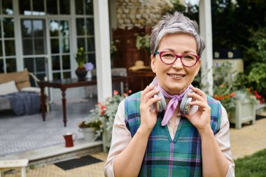 Şık gözlüklü neşeli olgun kadın kulaklıkla poz veriyor ve kameraya gülümsüyor.