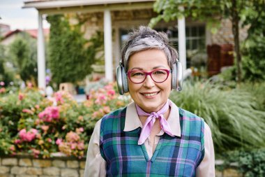 debonair cheerful mature woman in stylish glasses posing with headphones and smiling at camera clipart