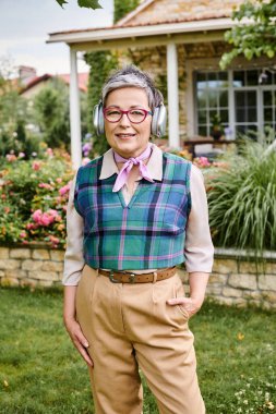 Şık gözlüklü neşeli olgun kadın kulaklıkla poz veriyor ve kameraya gülümsüyor.