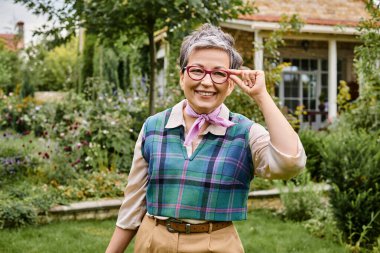sofistike, olgun, neşeli, kısa saçlı kadın İngiltere 'deki evinin yakınındaki kameraya gülümsüyor.