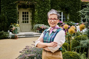 elegant mature joyous woman with short hair and glasses smiling at camera near her house in England clipart