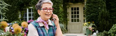 elegant cheerful mature woman with glasses talking by phone net to her house in England, banner clipart