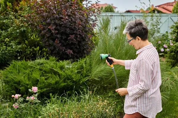 stock image good looking cheerful mature woman with glasses using hose to water her lively plants in her garden