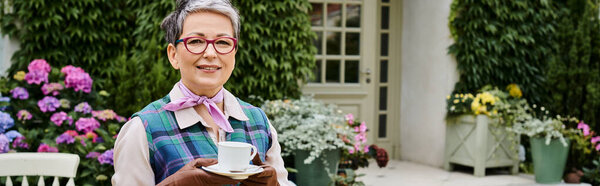 elegant jolly mature woman drinking tea near house in England and smiling at camera, banner