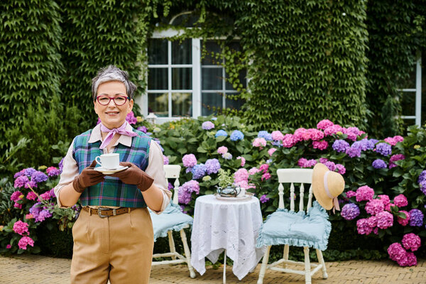 elegant jolly mature woman in chic attire drinking tea near house in England and smiling at camera