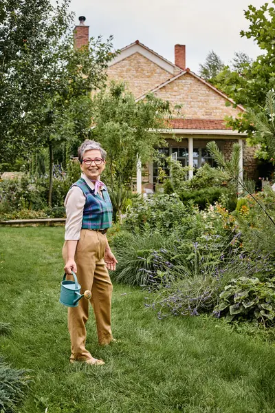 Şık, neşeli, olgun bir kadın. Sofistike giyinmiş. Elinde su kovası, kameraya gülümsüyor.