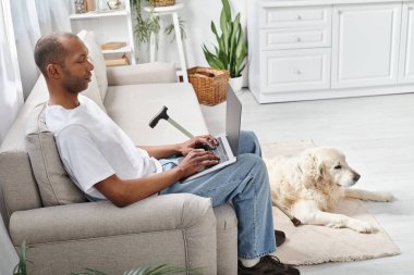 Miyastenia gravis 'i olan Afrikalı bir Amerikalı kanepeye oturmuş Labrador köpeğinin yanında dizüstü bilgisayar kullanıyor..