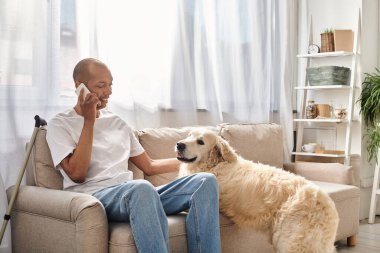 Miyastenia gravis 'i olan Afrikalı Amerikalı bir adam koltukta oturmuş sadık Labrador köpeğinin yanında telefonla konuşuyor..
