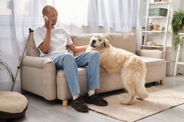Afrikalı Amerikalı bir adam evinde sadık Labrador köpeğinin yanında bir koltukta oturup cep telefonuyla konuşuyor..