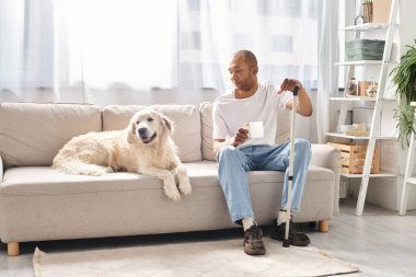 Miyastenia gravis 'i olan Afro-Amerikalı bir adam yanında Labrador köpeği ile kanepede oturuyor..