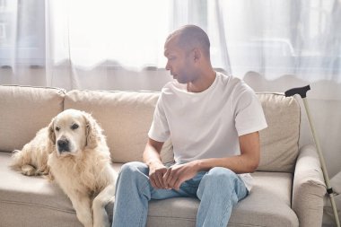 Özürlü bir Afro-Amerikan adam, miastenya gravis 'i olan sadık Labrador köpeğinin yanındaki kanepede dinleniyor..
