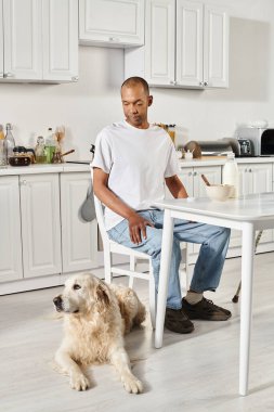 Engelli bir Afro-Amerikan adam mutfak masasında Labrador köpeğinin yanında oturuyor ve içten bir bağ kuruyor..