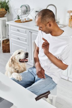 An African American man in a wheelchair and his loyal Labrador retriever enjoying a moment together in the kitchen. clipart